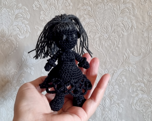 Nahaufnahme von einer gehäkelten Figur. Sie ist fast komplett aus schwarzer Wolle gemacht, nur die langen Haare und die Augen sind dunkelgrau.