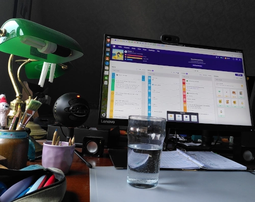 Photo von einem Schreibtisch, links eine Lampe mit einem grünen Glasschirm, rechts ein Computerbildschirm, davor ein großes Glas Wasser