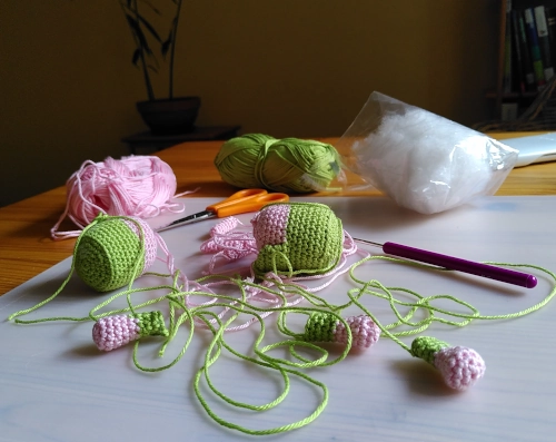 Gehäkelte Teile in rosa und grün, eine Häkelnadel und eine Tüte Füllwatte auf einem Tisch.