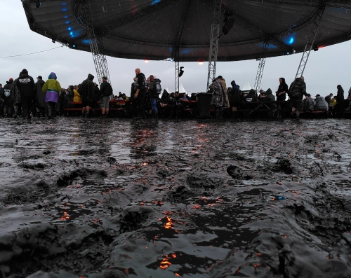 Matsch in Bodennnähe fotografiert, im Hintergrund ein riesiges Pavilliondach mit mehreren Menschen und Bänken darunter. Der Himmel ist grau.