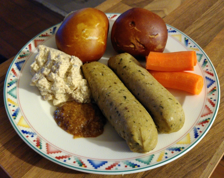 Ein Porzellanteller, darauf zwei helle vegane Würstchen, Karottensticks, zwei kleine Laugenbrötchen, Kartoffelsalat und ein Klecks süßer Senf.