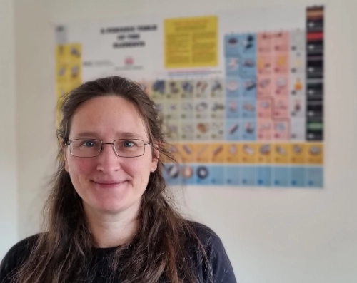 Nahhaufnahme einer Frau, im Hintergrund unscharf ein Periodensystem als Wandposter.