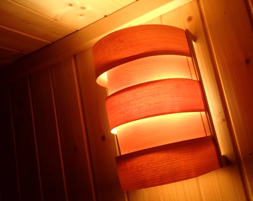 Nahaufnahme von einer Sauna-Wandlampe. Die Lampe leuchtet in der ansonsten eher dunklen Sauna. Dadurch, dass vor dem Leuchtmittel dünne Holzstreifen sind, ist das Licht orangefarben und warm.