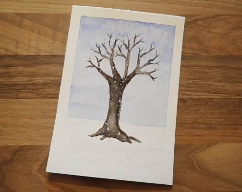 Ein circa DINA5 großes Stück Aquarellpapier, darauf gemalt ein unbelaubter Baum vor einem hellblauen Himmel, im Vordergrund viele weiße Tupfen, um Schneeflocken darzustellen.