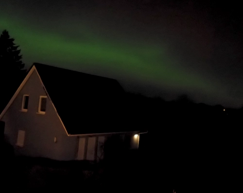 Ein eEnfamilienhaus bei Nacht aus einem Dachfenster fotografiert, am Himmel darüber ein leuchtend grüner Streifen.