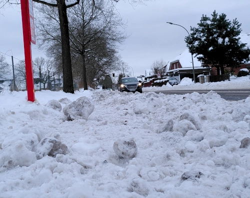 Aufnahme in Bodennähe von einer Bushaltestelle. Der Fußweg ist überschüttet mit Schneebrocken und Eisklumpen, zur Straße hin ist ein mehr als knöchelhoher Wall aus Schnee.