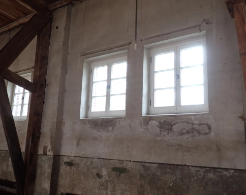 Eine Wand in einer Halle, mit Balken verstärkt. Eine Reihe von Fenstern lässt nur den leuchtend grauen Himmel draußen sehen.