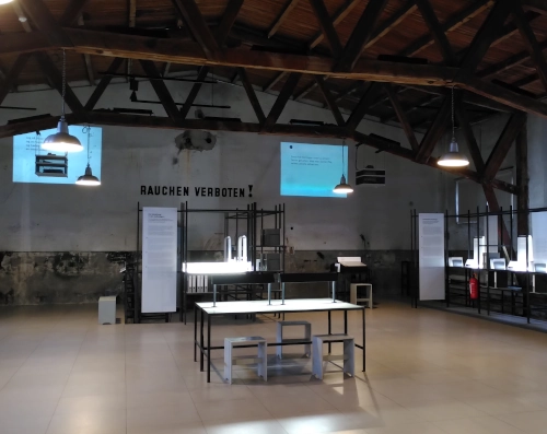 Blick in einen Museumsraum mit Balken an der Decke, Tischen mit Informationsmaterial, Projektionen an der Hinterwand und mehreren Stationen mit Kopfhörern. An der hinteren Wand steht "Rauchen verboten", da es sich hierbei um eine ehemalige Garage im KZ Neuengamme handelt.