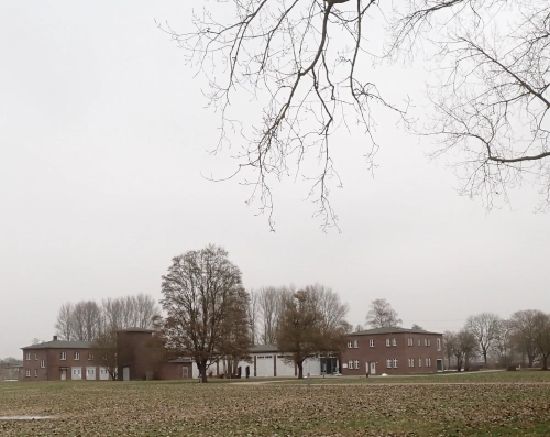 Eine flache, offene Landschaft, im Hintergrund Baracken, zum Teil aus roten Klinkerziegeln.