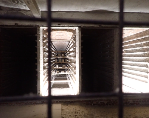 Blick in zwei hell erleuchtete Räume mit horizontalen Schienen aus Steinen an den Seitenwänden. In diese sind Tafeln hinein geschoben, auf denen einzelne Ziegelsteine stehen. Der  Vordergrund ist dunkel und mit einem Metallgitter abgesperrt, das durch die Kameraeinstellung unscharf erscheint.