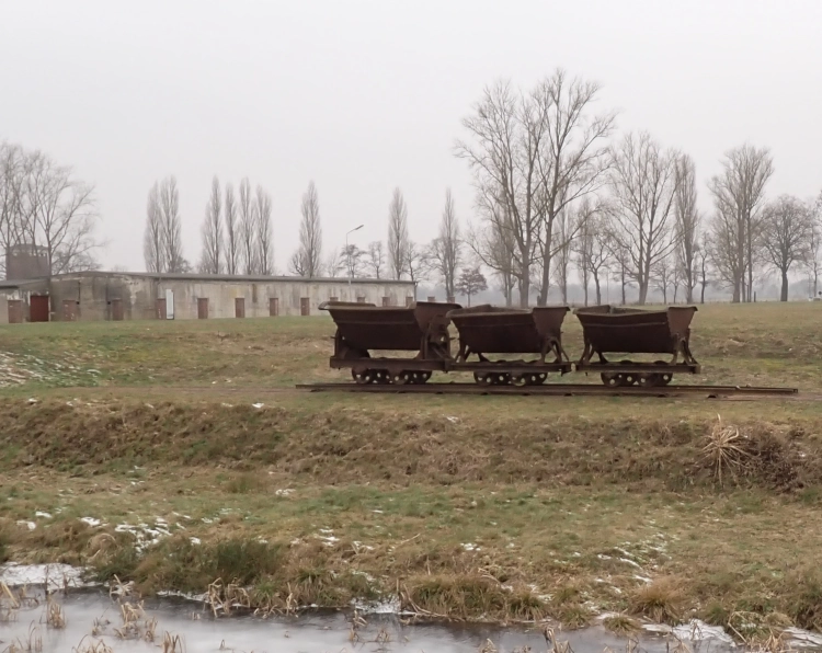 Außenaufnahme in der Gedenkstätte Neuengamme, drei Loren auf einer Schiene, im Vordergrund ein zugefrorener Graben, Im Hintergrund unbelaubte Bäume, Baracken und grauer Himmel