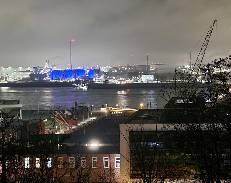 Der Containerhafen in Hamburg bei Nacht. Der Himmel hängt voller dunkelgrauer Wolken, davor glitzern viele weiße Lichter. Ein Gebäude leuchtet allerdings blau.