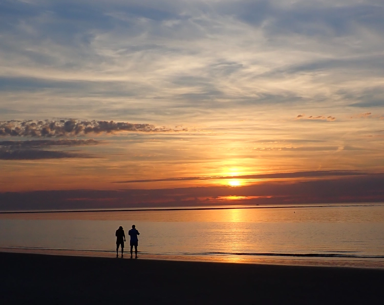 Ein farbenfroher Sonnenuntergang am Meer, der Himmel ist von leichten Wolken durchzogen. Am Strand stehen zwei Personen, nur als Umrisse sichtbar.