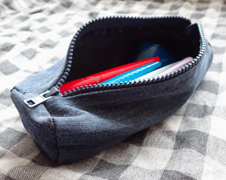 Auf grau-schwarz kariertem Stoff liegt eine geöffnete Federmappe aus blauem Jeansstoff, darin ein paar Füller in vielen unterschiedlichen Farben.