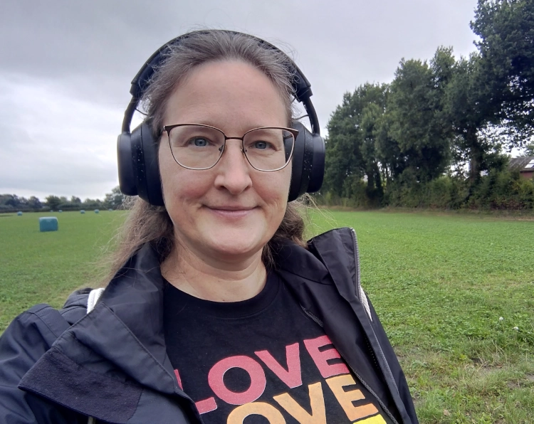 Eine Frau in Regenjacke steht in einer Landschaft und trägt einen Kopfhörer, mit dem sie ihre Hörbuchapp hört