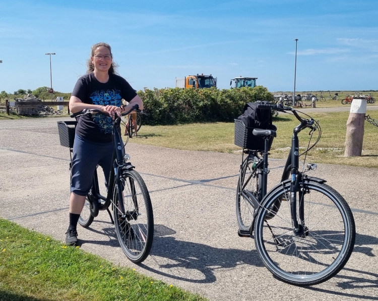 Eine Frau steht sommerlich gekleidet mit einem Fahrrad vor einer Inselhafenanlage. Neben ihr lehnt ein Fahrrad ohne Fahrer auf dem Ständer. Die Frau lächelt in die Kamera.