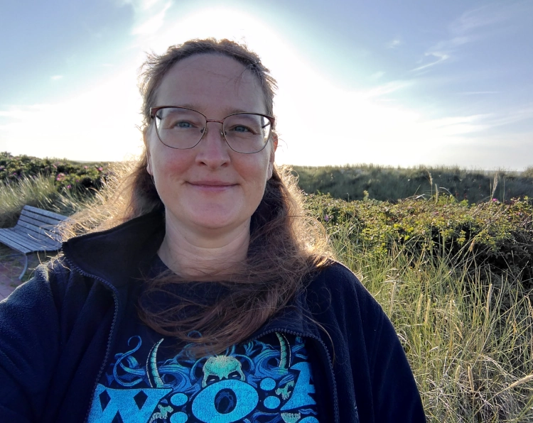 Eine Frau steht vor Dünen, die Sonne beleuchtet sie von hinten. Sie trägt ein T-Shirt, eine Fleecejacke und ein GRinden.
