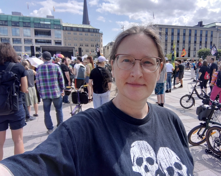 Eine Frau auf einem öffentlichen Platz. Sie trägt ein schwarzes Shirt mit zwei Piratenschädeln. Hinter ihr sind weitere Teilnehmer:innen einer Demonstration.