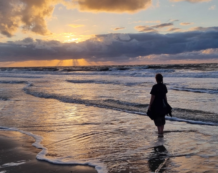 Eine Frau steht bei einem farbenprächtigen Sonnenuntergang am Strand, mit den Füßen im Wasser und das Gewicht zur Sonne hin gedret. Sie ist nur als Umriss zu sehen und hat offenbar eine Jacke um die Hüften gebunden.