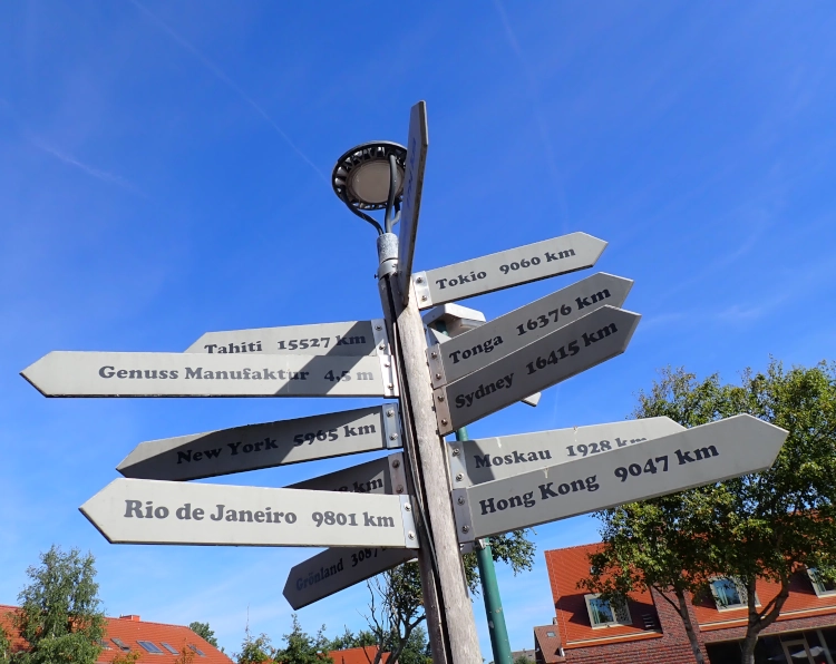 Ein Wegweise gegen einen blauen Himmel abgebildet. Die Ziele sind teils international, zum Beispiel Tokio, Moskau, Tahiti und Rio de Janeiro. Eins der Ziele ist aber die Genussmanufaktur in 4,5 Metern Entfernung.