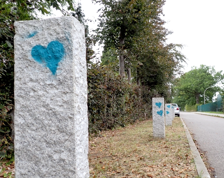 Granitpfosten an einem Straßenrand. Das Gras um die Pfosten herum ist trocken und gelb, auf die Steine hat jemand leuchtend blaue Herzen gesprüht.