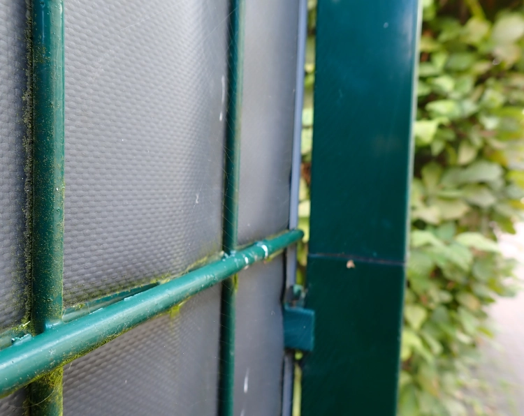 Im Vordergrund nah aufgenommen ein dunkelgrüner Metallzaun mit einer grauen Kunststoffplane, im Hintergrund unscharf eine grün belaubte Buchenhecke.