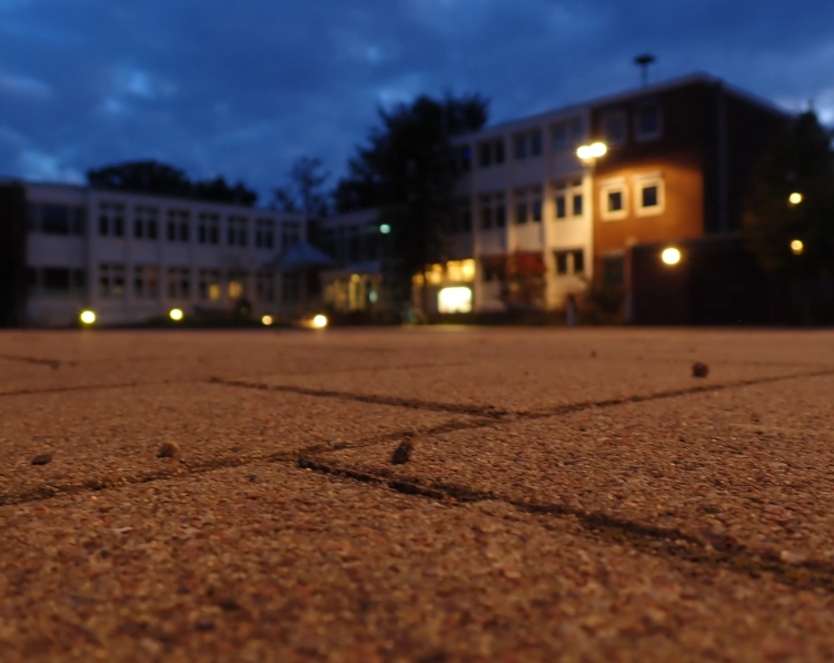 Ein mit Laternen beleuchtetes Verwaltungsgebäude aus roten Steinen mit weißen Elementen steht unscharf im Hintergrund vor einem bewölkten Herbstabendhimmel. Im Vordergrund sind Pflastersteine von Erdboden aus aufgenommen im Fokus.